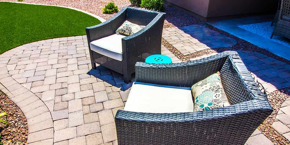Wicker patio chairs with cushions on a paved courtyard, adjacent to artificial turf, creating a cozy outdoor seating area.