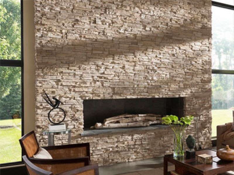 A spacious living room with a large stone fireplace wall. The fireplace has stacked wood and a decorative sculpture on the mantel. Large windows on either side allow natural light to fill the room, highlighting a small table with a plant.