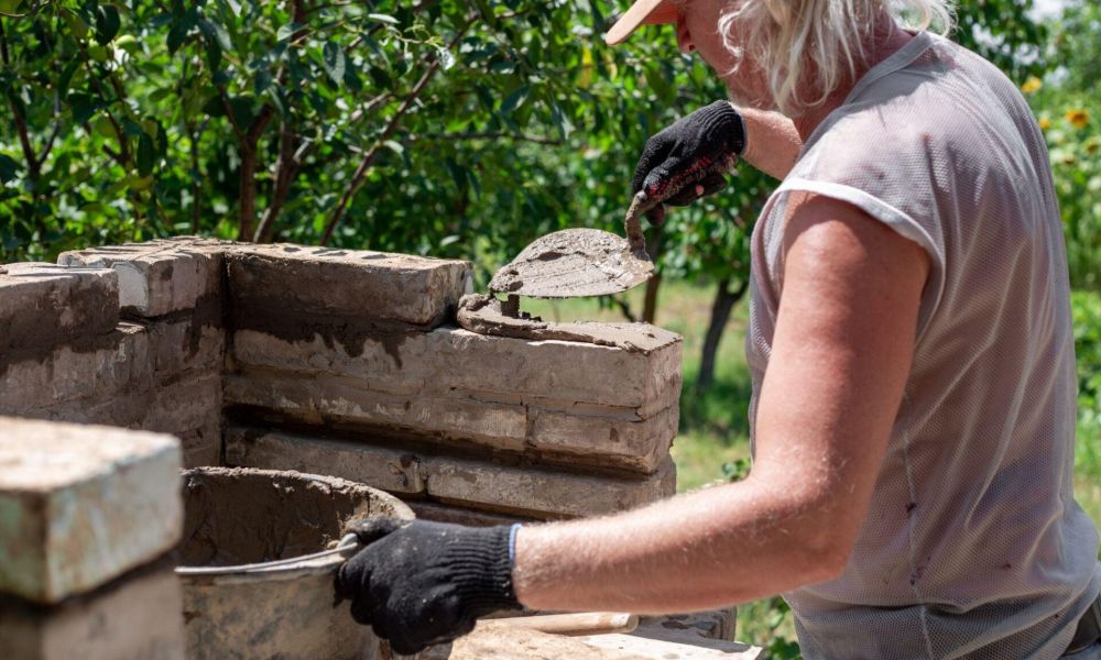 Person building a brick wall with mortar in a garden setting, wearing a cap and gloves.