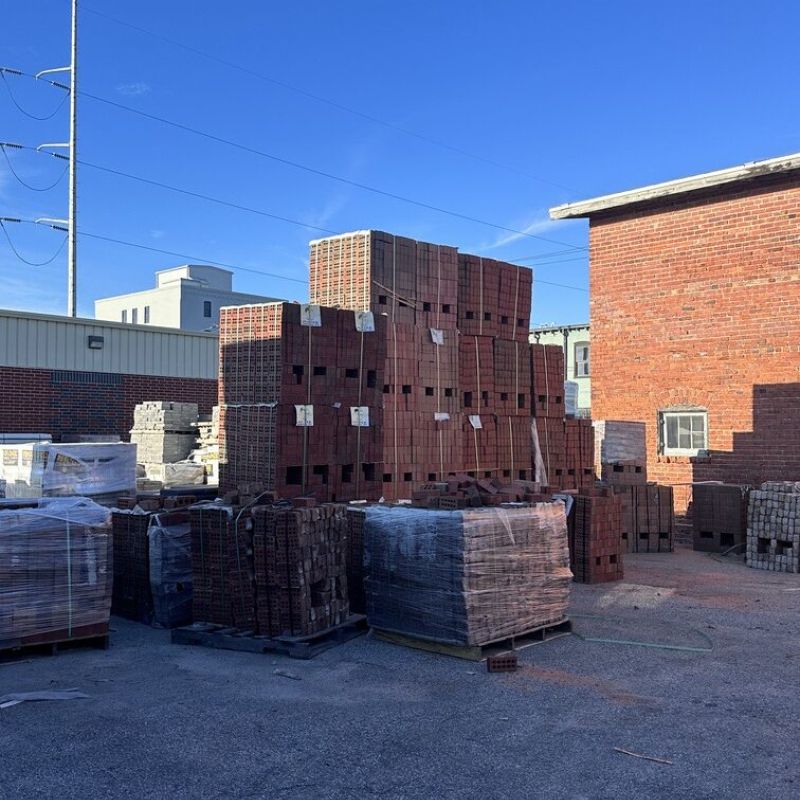 A stack of red clay bricks and other building materials are arranged outdoors on a concrete surface. Nearby are additional stacks covered by plastic wrap. The background includes industrial buildings, a brick wall, and a clear blue sky.