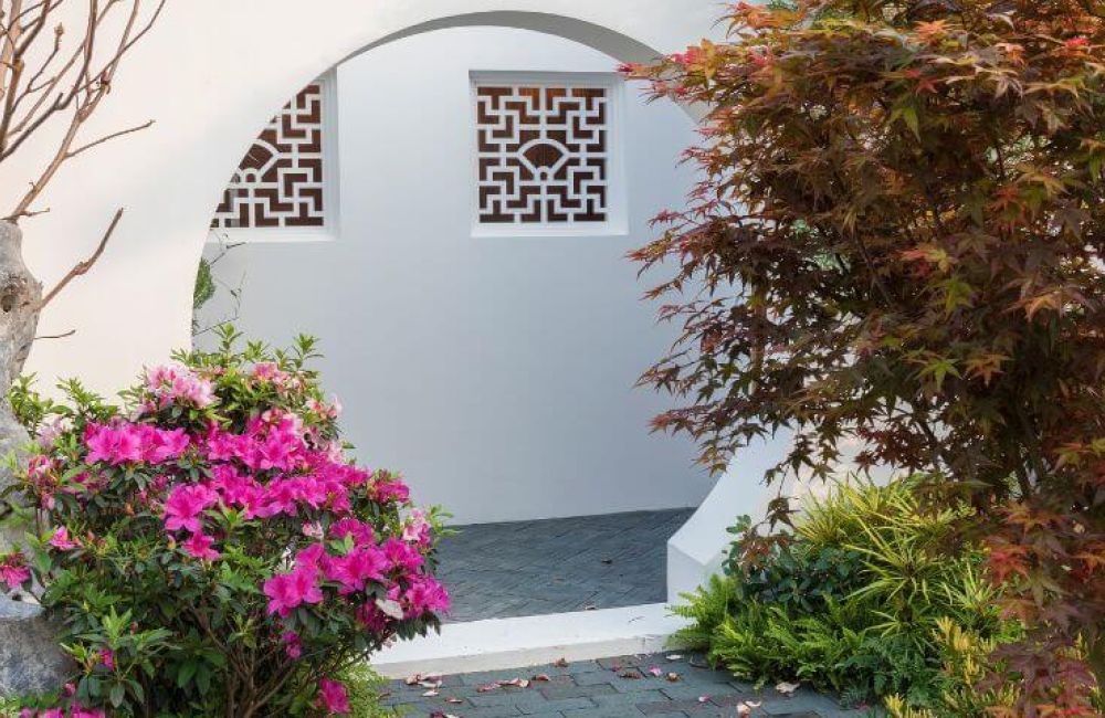 A garden with pink flowers and a stone, framed by a circular arch. Ornate windows sit in a white wall. Autumnal leaves add color, enhancing the serene, Asian-inspired setting.