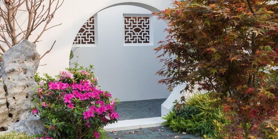 A garden with pink flowers and a stone, framed by a circular arch. Ornate windows sit in a white wall. Autumnal leaves add color, enhancing the serene, Asian-inspired setting.