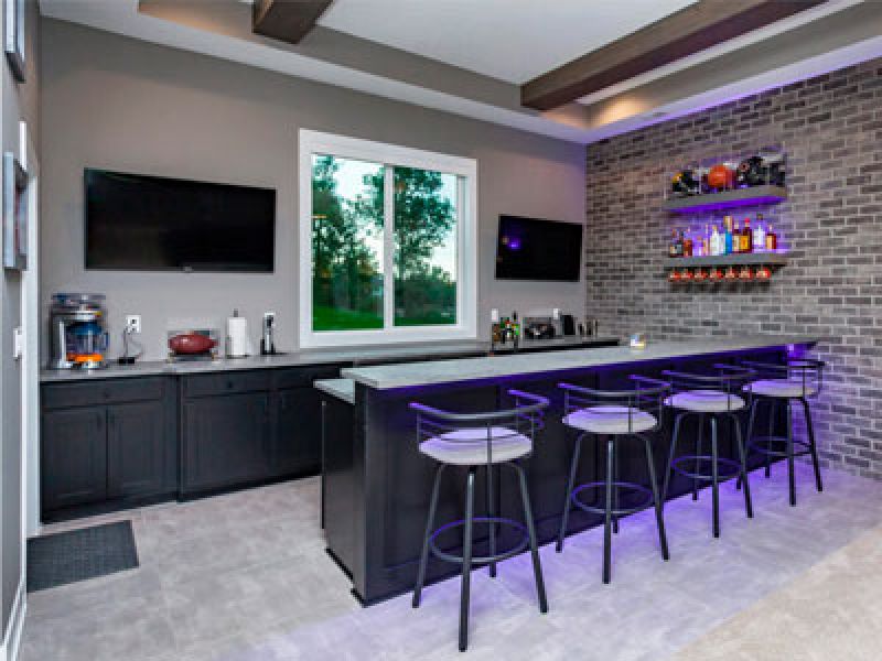 A modern home bar with a sleek counter lined with four stools under soft purple lighting. Brick accent wall features shelves with bottles and glasses. Two wall-mounted TVs and a large window with a greenery view complete the space.