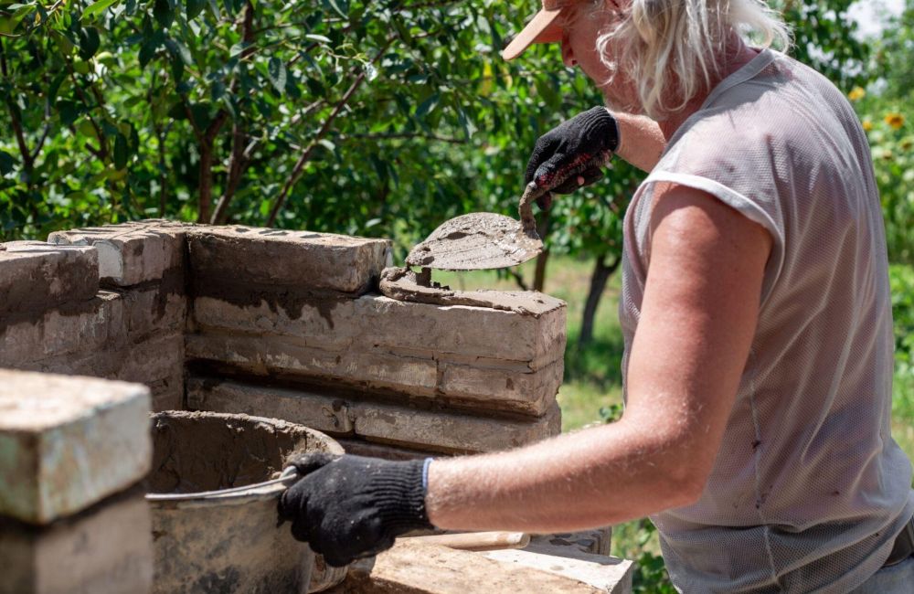 Person building a brick wall with mortar in a garden setting, wearing a cap and gloves.