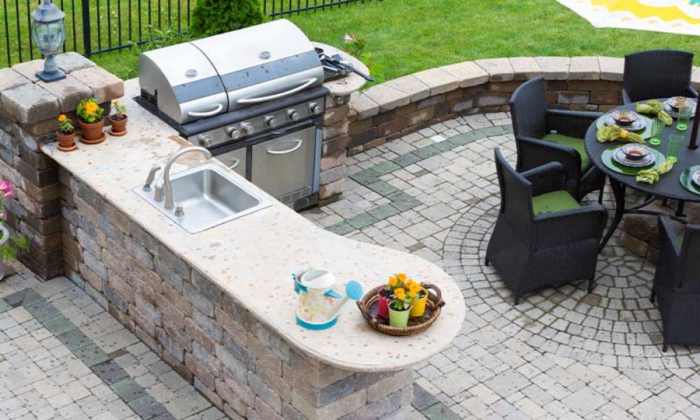 Outdoor kitchen with grill, sink, and patio dining area with round table and umbrella, surrounded by lush green grass.