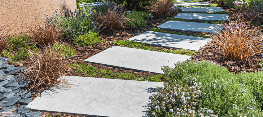 A garden path with large rectangular stone slabs leads through lush greenery, including shrubs and ornamental grasses. The path is bordered by small rocks and surrounded by various plants, creating a serene and inviting scene.
