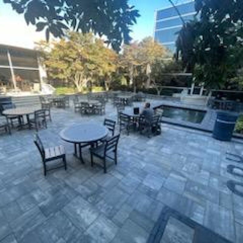A spacious outdoor patio with several empty tables and chairs surrounded by trees and greenery. A person sits at a table working on a laptop. A building is visible in the background under a clear blue sky.