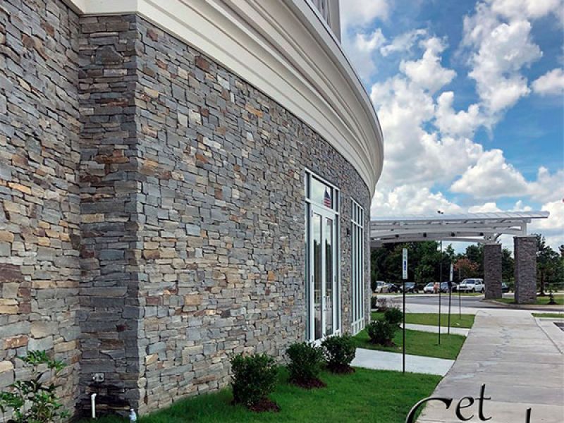 Modern building exterior with stone veneer walls and large windows. A curved pathway runs alongside, bordered by greenery. The sky is partly cloudy. The image is labeled Get Real Stone in the bottom right corner.