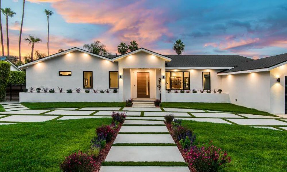 A modern single-story house stands at dusk, with geometric lawn patterns made of hardscapes and decorative shrubbery under a colorful sky.