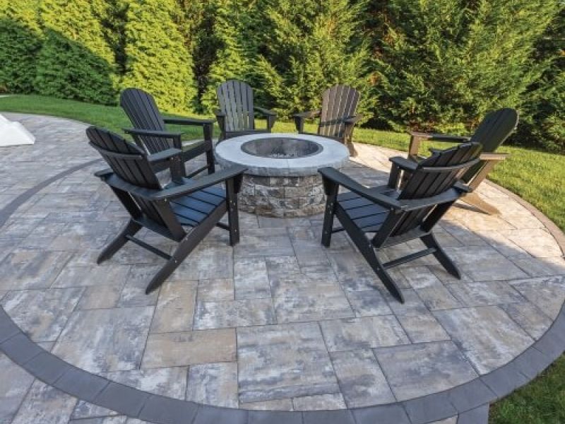 Patio with fire pit and Adirondack chairs, set on stone pavers amidst a lush green garden backdrop.