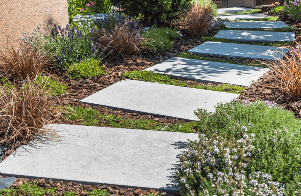 A garden path with large rectangular stone slabs leads through lush greenery, including shrubs and ornamental grasses. The path is bordered by small rocks and surrounded by various plants, creating a serene and inviting scene.