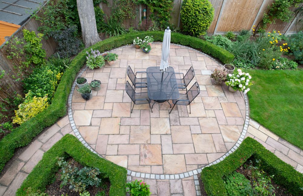 Circular patio garden with stone tiles and outdoor dining set, surrounded by lush greenery and flowers.