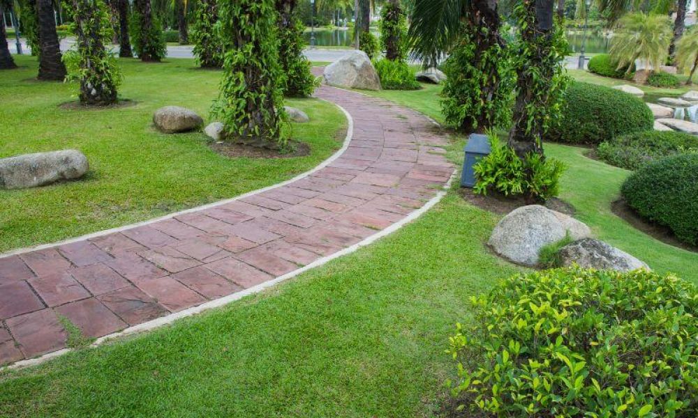 A winding red brick pathway meanders through a lush green park with manicured shrubs, palm trees, and interspersed large rocks.