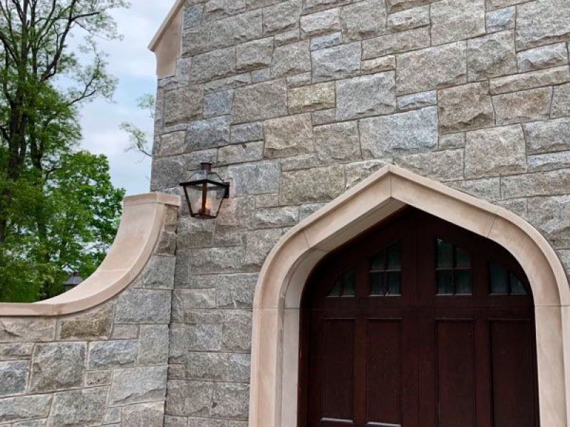 A stone building with a wooden arched door and a lantern mounted on the wall. The structure features beige stone blocks and has a small section of wall curving upward. Trees are visible in the background under a cloudy sky.