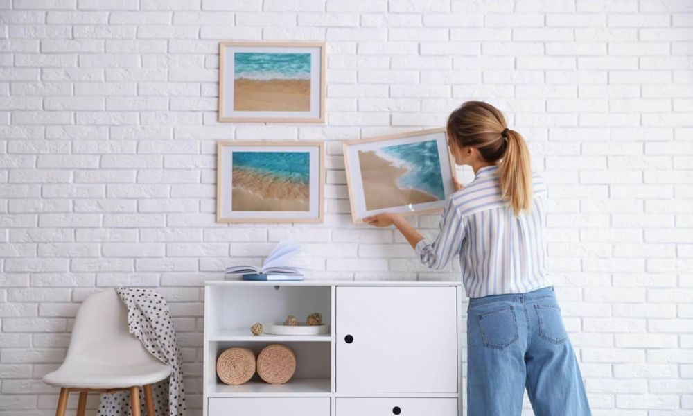 Woman arranging framed ocean artworks on a white brick wall for modern home decor.