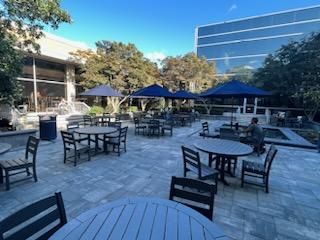 Outdoor patio with several round wooden tables and chairs under blue umbrellas. Few people are seated, surrounded by modern buildings and greenery. The sky is clear and sunny, creating a relaxing atmosphere.