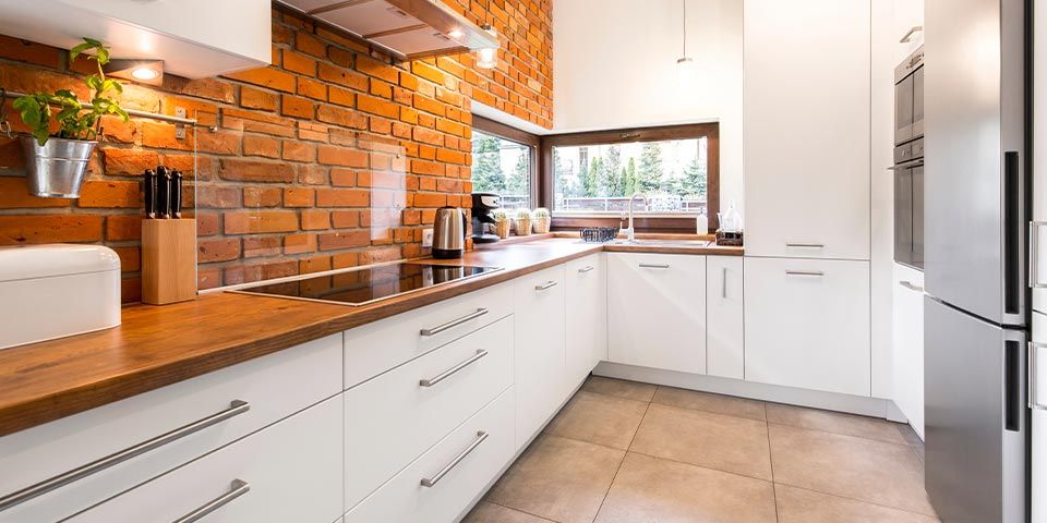 Modern kitchen with white cabinets, wood countertops, brick wall, large window, and stainless steel appliances.