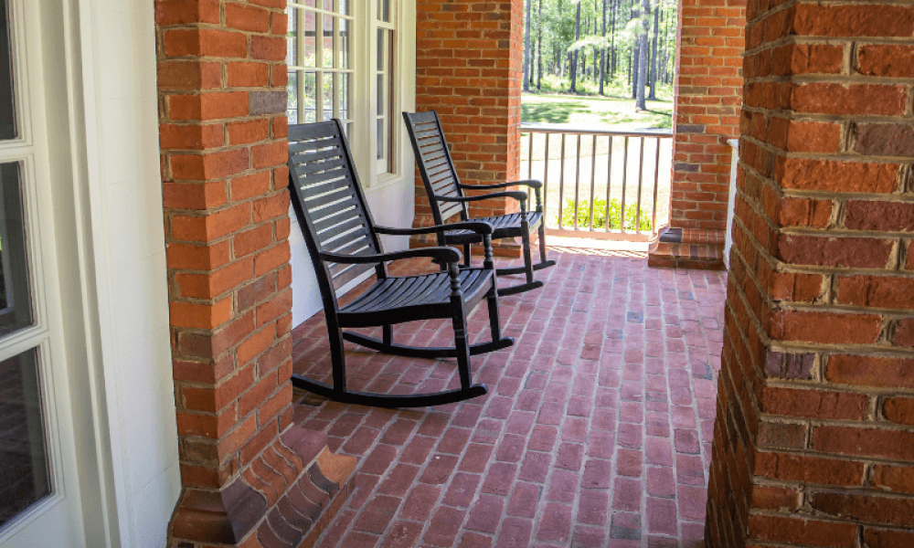 Two black rocking chairs sit on a brick porch, surrounded by red brick walls and overlooking a lawn with trees in the background.