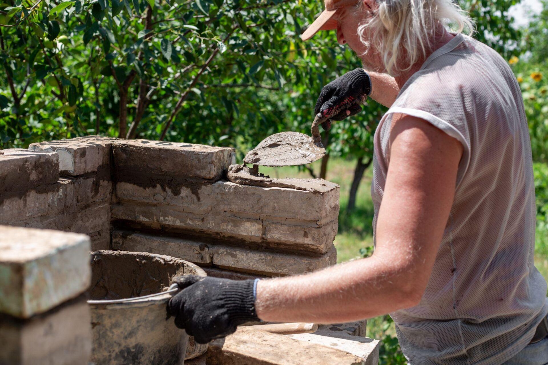 Person building a brick wall with mortar in a garden setting, wearing a cap and gloves.