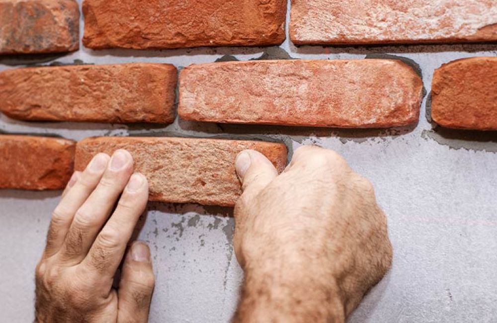 Hands installing red brick veneer on a wall for home construction or renovation.