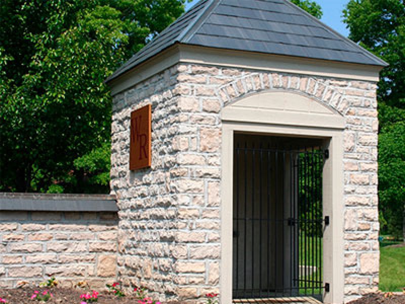 A small stone structure with a pointed roof and iron gate, set against a green leafy background. A sign with TR is mounted on the wall. Pink flowers and shrubs are near the base.