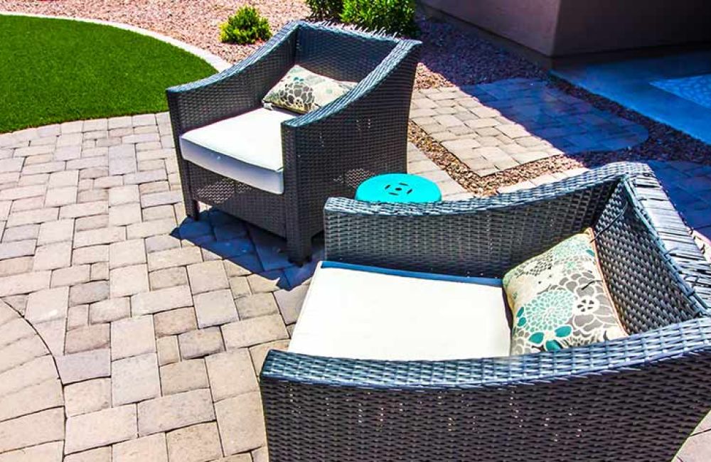 Wicker patio chairs with cushions on a paved courtyard, adjacent to artificial turf, creating a cozy outdoor seating area.