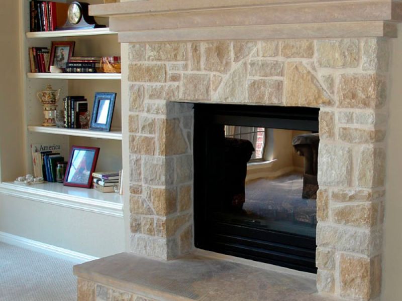 Stone fireplace with a rectangular opening surrounded by light brown stone bricks. Built-in shelves on the left hold books and decorative items. A carpeted floor leads to the fireplace, which reflects a cozy seating area in its glass pane.
