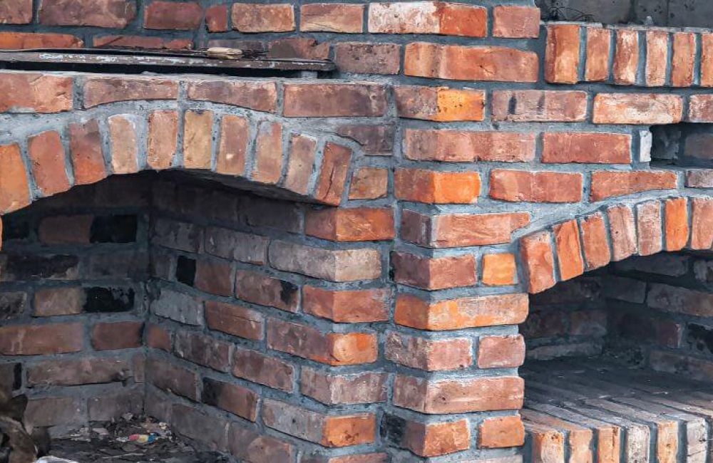 Brick oven structure with arched openings; located outdoors, the red and brown bricks form two separate oven chambers, set against a rustic environment with visible mortar joints.