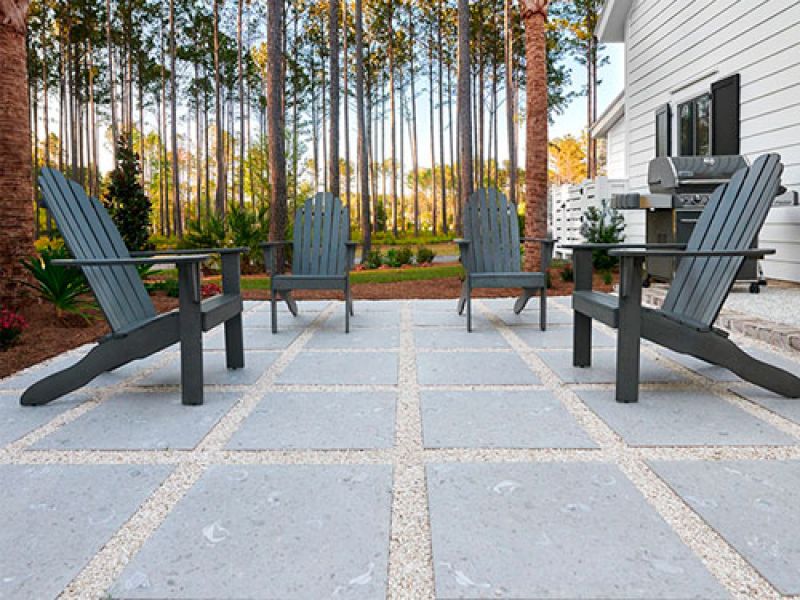 Patio scene with four dark wooden Adirondack chairs on a tiled surface, surrounded by trees. A barbecue grill is visible on the right, near a white house wall. The setting suggests a peaceful outdoor gathering space.