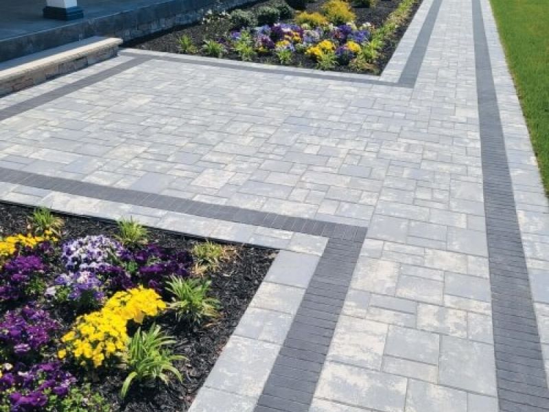 Modern garden pathway with gray pavers, vibrant flowers, and greenery next to a spacious lawn and patio area.