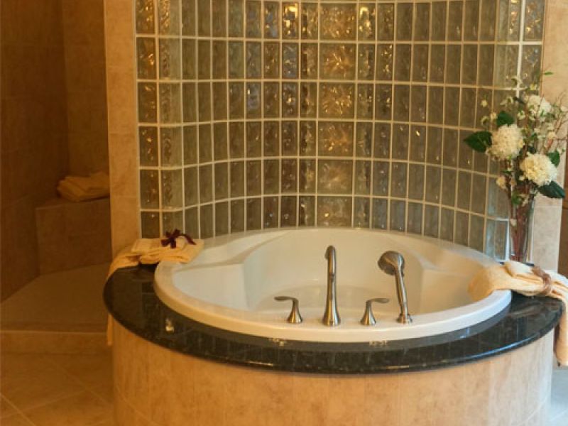 A luxurious bathroom features a large, round bathtub with silver fixtures set against a wall of glass tiles. Towels are neatly placed on the tubs edge, and a floral arrangement sits nearby on the tiled floor.