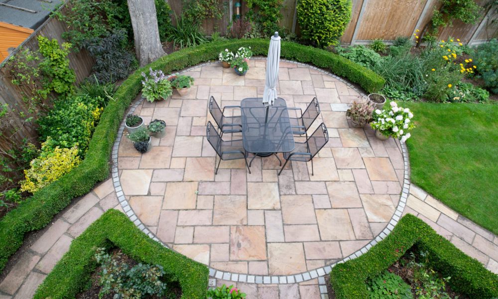 Circular patio garden with stone tiles and outdoor dining set, surrounded by lush greenery and flowers.
