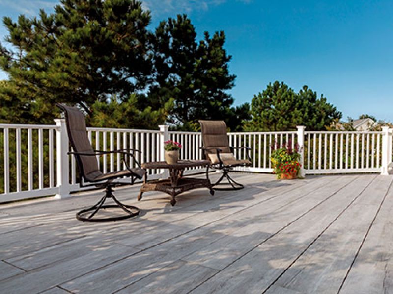 A spacious wooden deck with two wicker chairs and a table, holding a potted plant. The deck is enclosed by a white railing, and lush green trees are visible in the background under a clear blue sky.