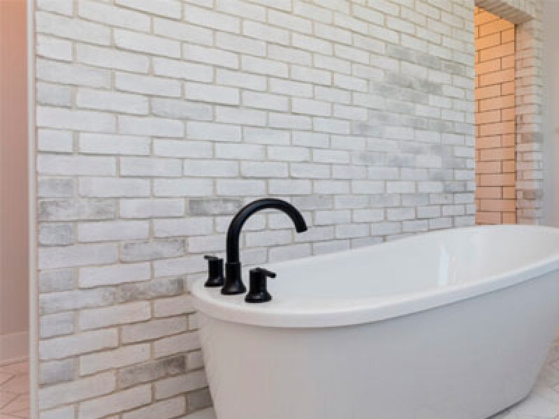 A modern bathroom features a white freestanding bathtub with black faucets, set against a backdrop of white brick walls. The room has a minimalist and stylish design.