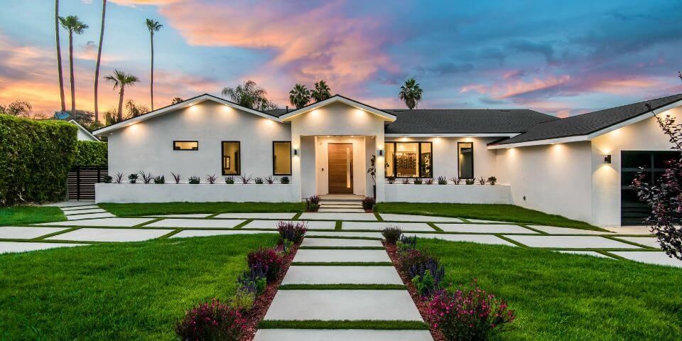 A modern single-story house stands at dusk, with geometric lawn patterns made of hardscapes and decorative shrubbery under a colorful sky.