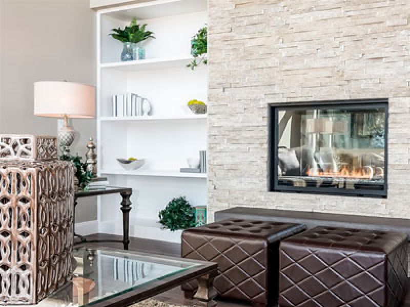 A modern living room with a stone fireplace, two brown ottomans, and a glass coffee table. A white bookshelf with plants and decor is on the left. A table lamp with a textured base is next to the bookshelf.