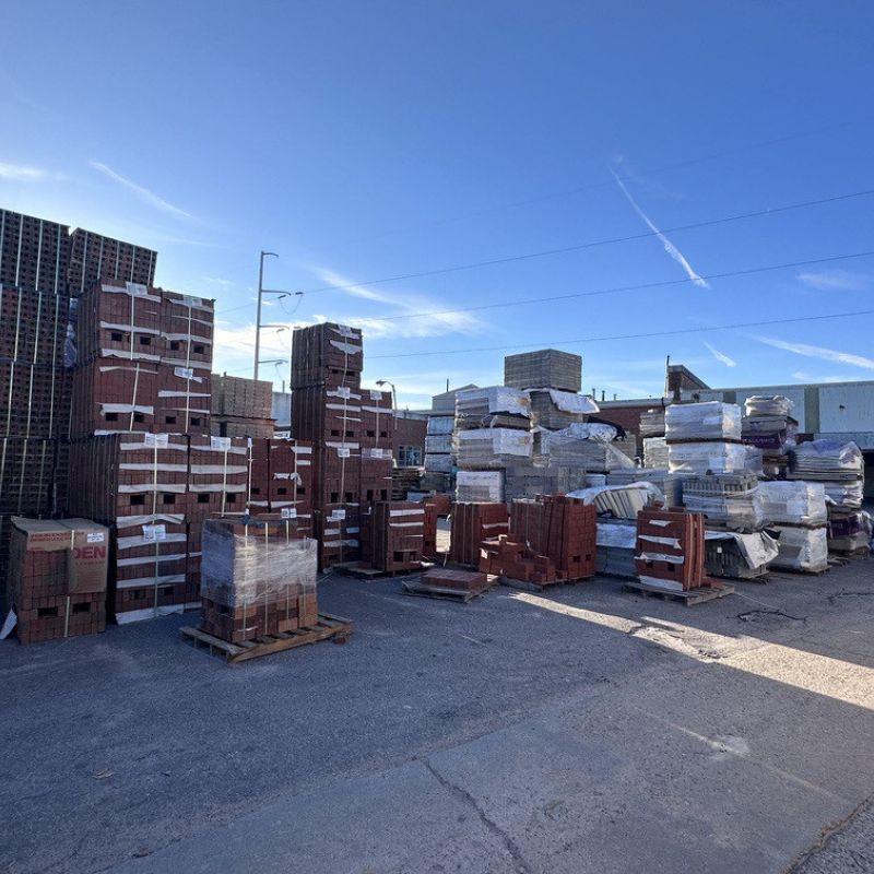A storage yard with stacks of bricks and building materials on wooden pallets. The sky is clear and blue, and there are power lines visible in the background. The ground is paved with asphalt.