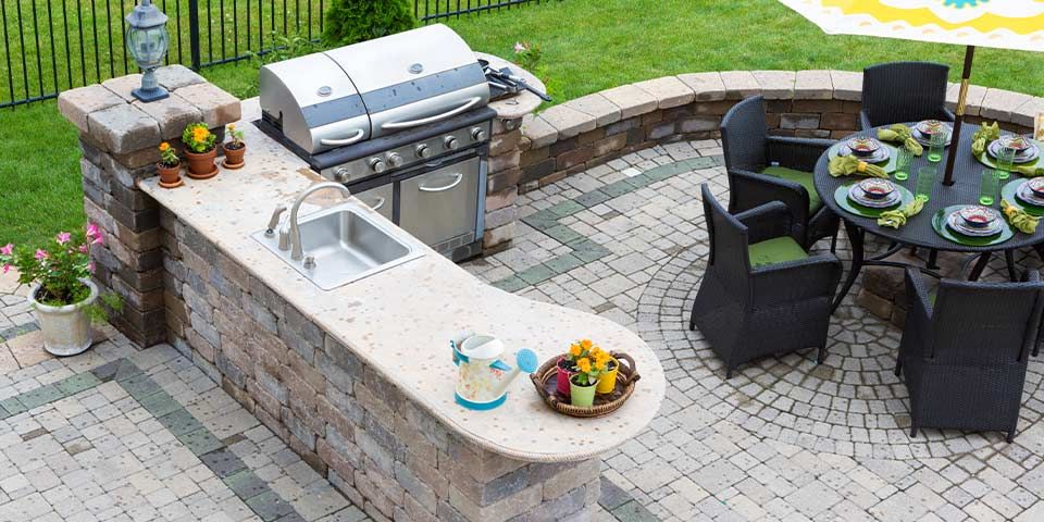 Outdoor kitchen with grill, sink, and patio dining area with round table and umbrella, surrounded by lush green grass.