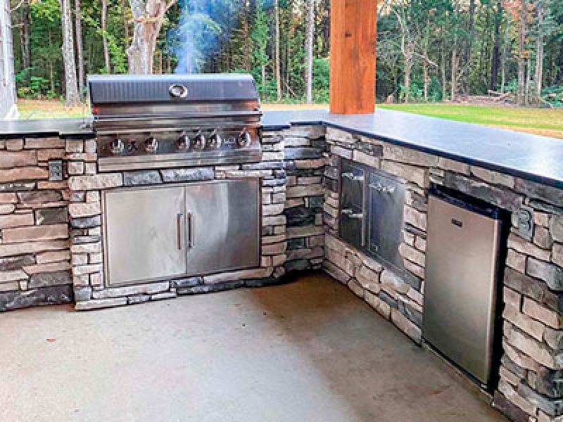 Outdoor kitchen with a stone veneer design featuring a stainless steel grill, smoker, cabinets, and a mini fridge. The area is under a wooden pergola, with a view of trees in the background. Smoke is rising from the grill.