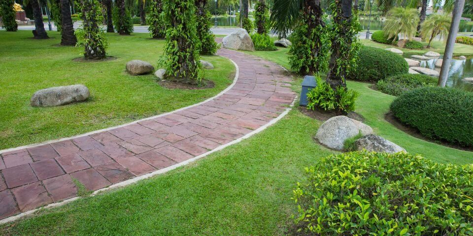A winding red brick pathway meanders through a lush green park with manicured shrubs, palm trees, and interspersed large rocks.