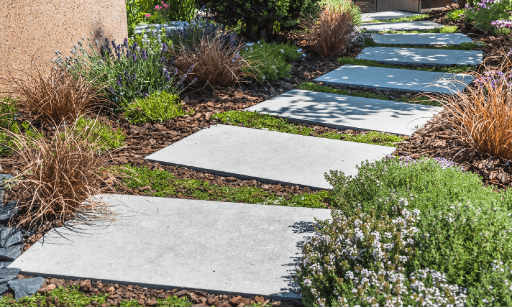 A garden path with large rectangular stone slabs leads through lush greenery, including shrubs and ornamental grasses. The path is bordered by small rocks and surrounded by various plants, creating a serene and inviting scene.