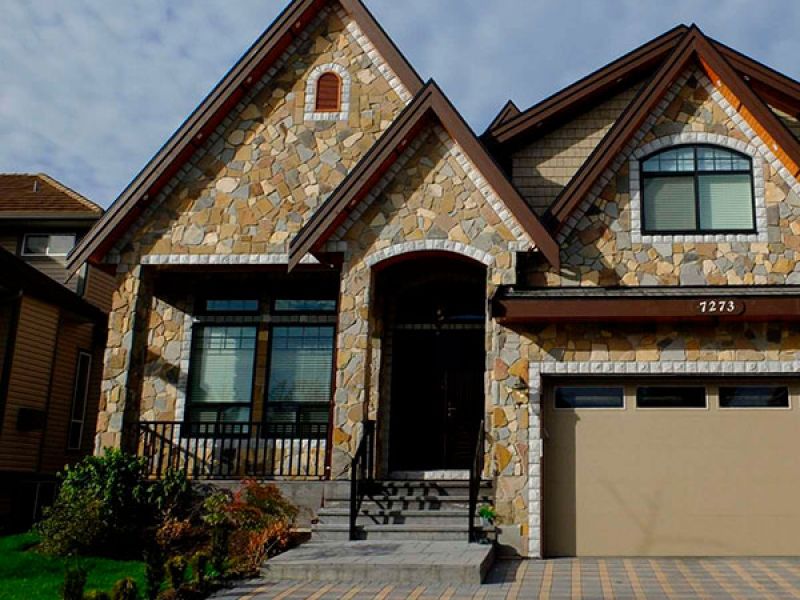 A two-story house with a stone facade, large windows, and a double garage. The driveway is paved with square tiles in a checkered pattern. Green bushes line the walkway leading to the front steps.