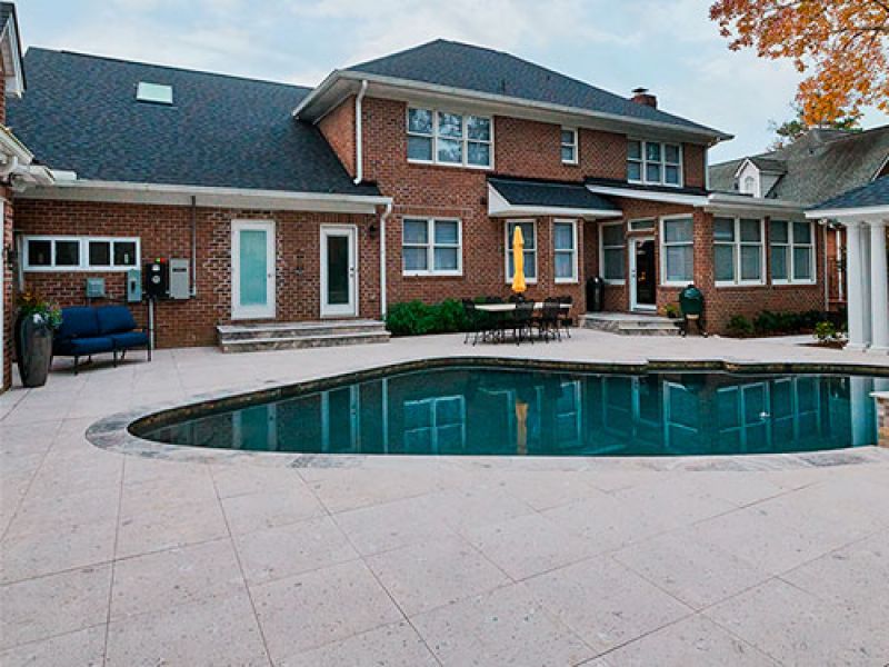 A spacious backyard features a large, kidney-shaped swimming pool surrounded by a concrete patio. Adjacent to the pool is a two-story brick house with multiple windows and doors. There is outdoor seating and a yellow umbrella nearby.