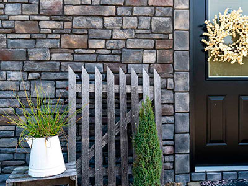 A rustic porch scene features a stone wall, a black door adorned with a light-colored wreath, and a small wooden fence gate. A white pitcher with grass sits on a bench next to a tall, slender evergreen plant.