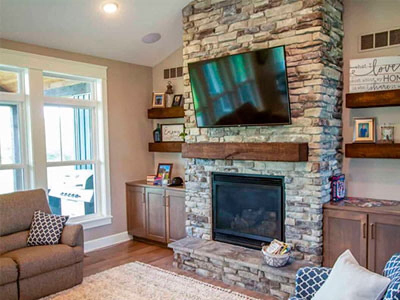 A cozy living room featuring a stone fireplace with a mounted TV above. Wooden shelves on either side display decor and photos. A large window lets in natural light. Theres a comfortable armchair and a basket beside the fireplace.