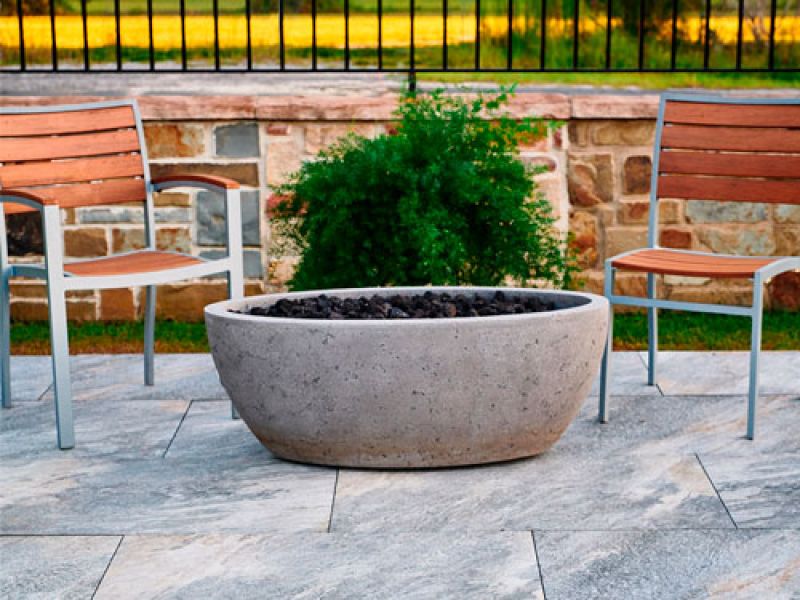 Outdoor scene with two wooden chairs and a large, round stone fire pit filled with dark rocks on a stone-tiled patio. A lush green plant and a stone wall are in the background, with a railing and grassy area beyond.