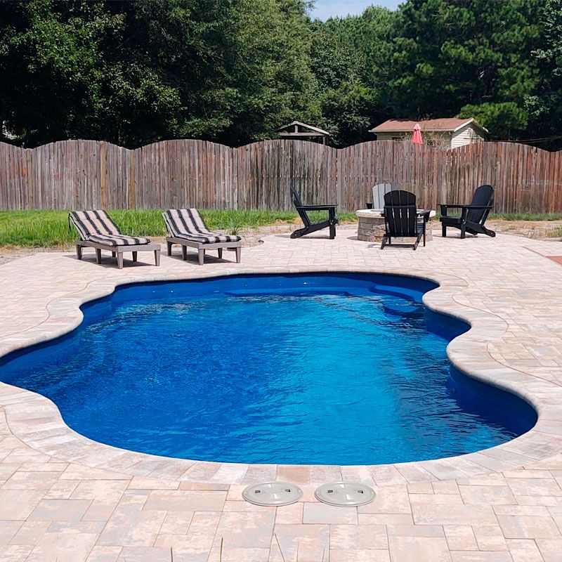 A backyard scene with a kidney-shaped swimming pool. Surrounding the pool are several lounge chairs with striped cushions and Adirondack chairs around a fire pit. A wooden fence encloses the area, and trees are visible in the background.