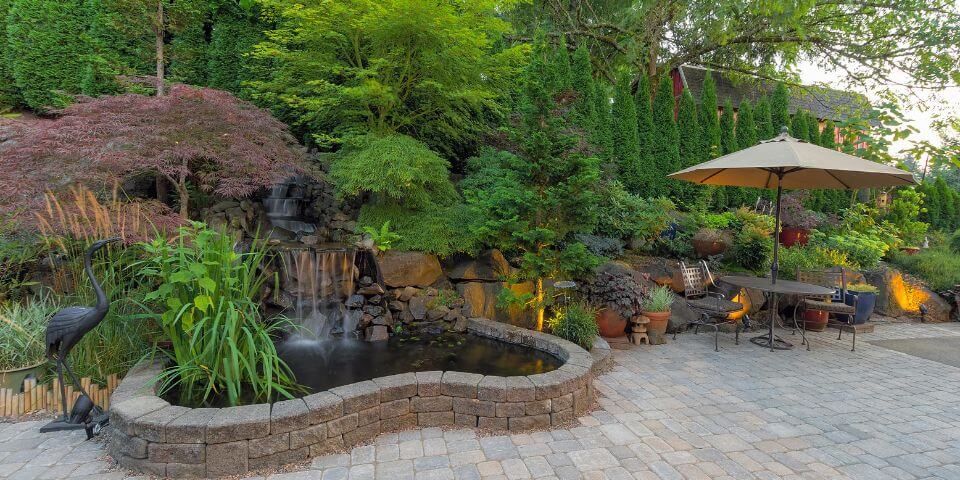 A landscaped garden with a waterfall, pond, and patio area featuring a fire pit, outdoor furniture, and an umbrella, surrounded by lush greenery and evening lighting.