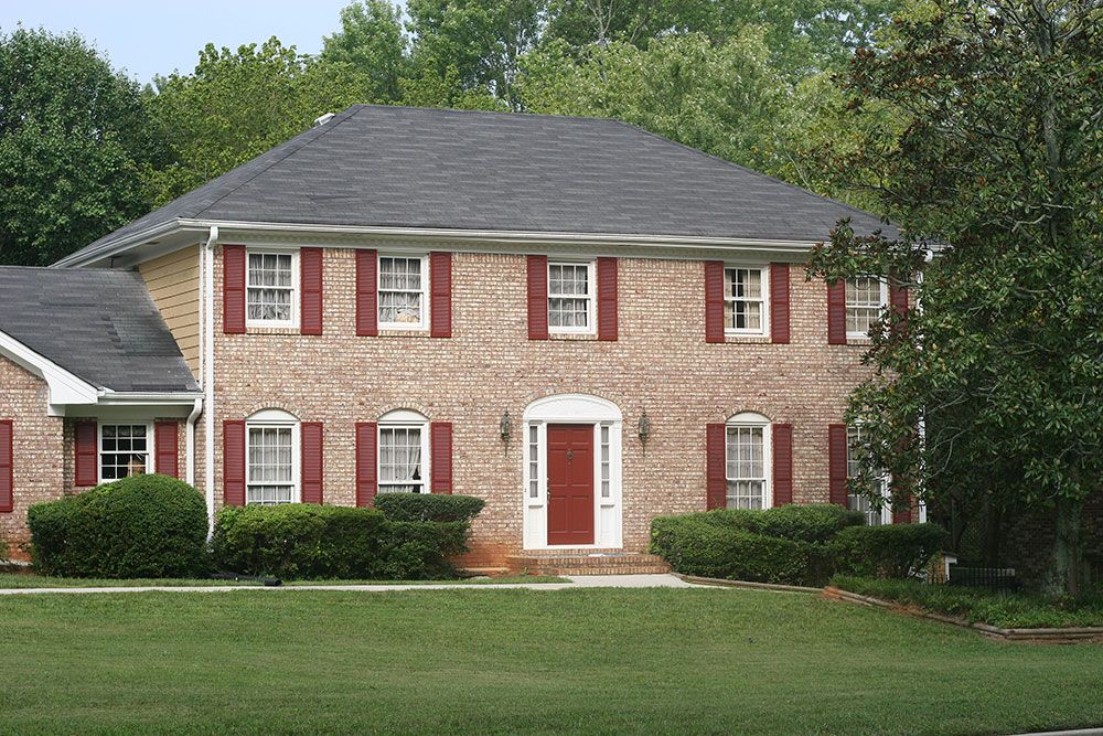 Two-story brick house with red shutters, a red door, and manicured shrubs, surrounded by green lawn and trees.