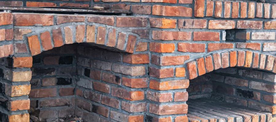 Brick oven structure with arched openings; located outdoors, the red and brown bricks form two separate oven chambers, set against a rustic environment with visible mortar joints.